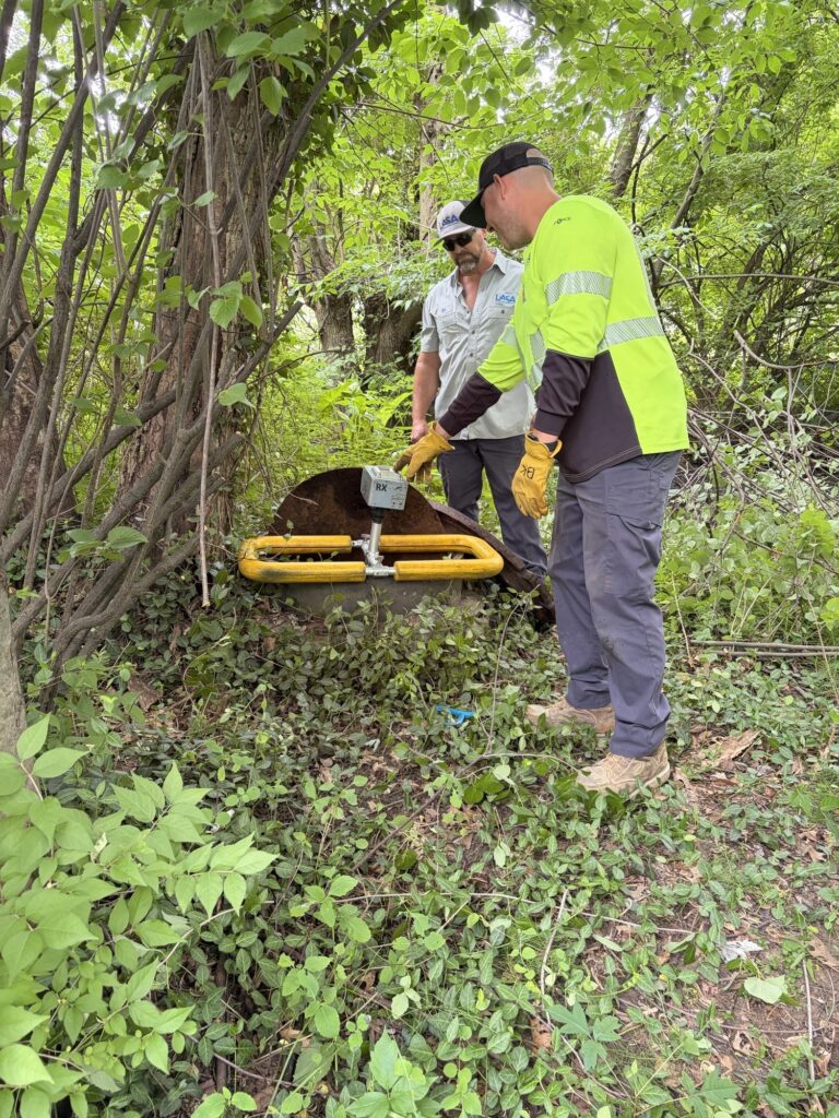 Two men inspecting an acoustic sewer screening device placed over a manhole in a wooded area.