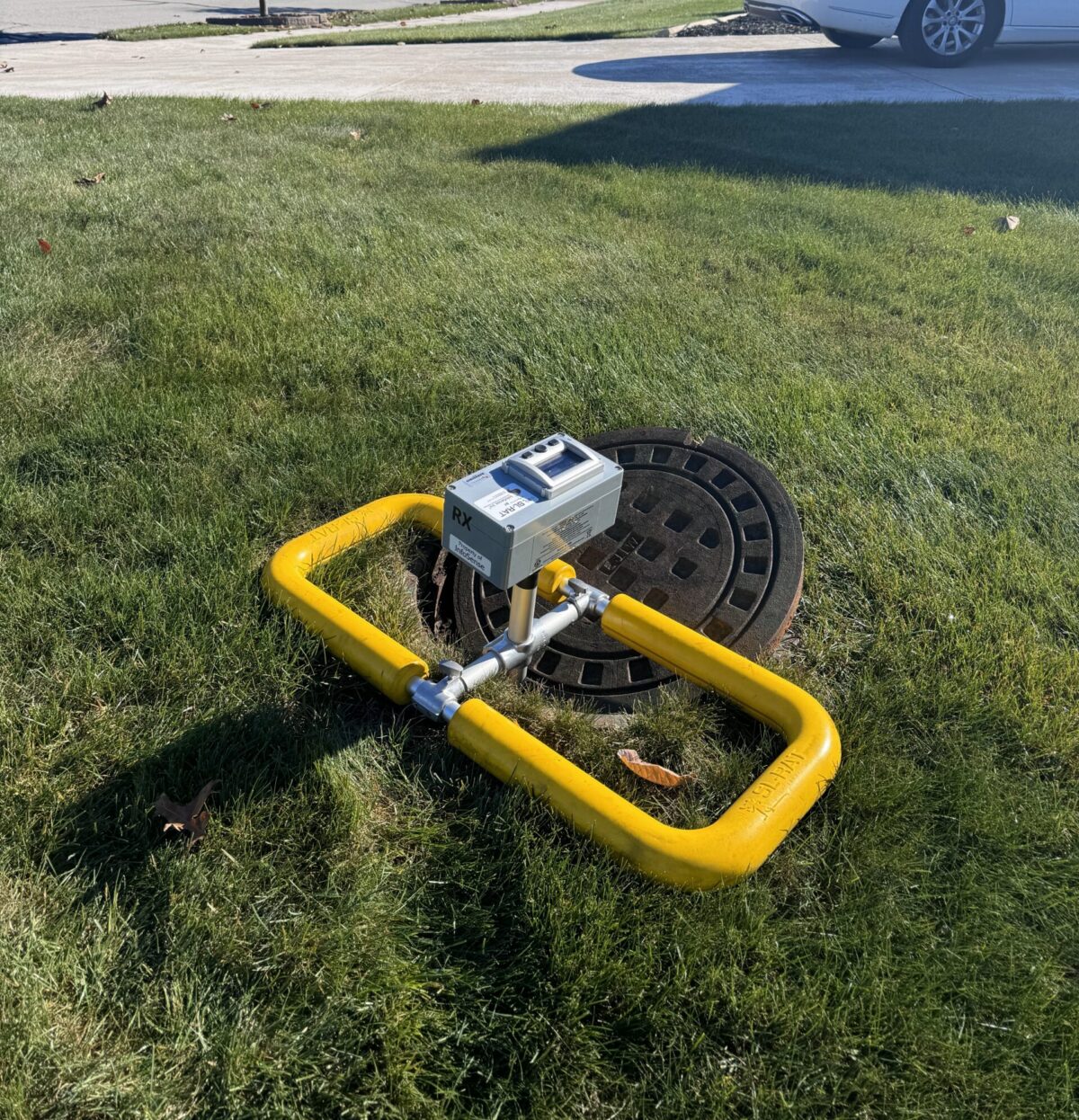 A SL-RAT unit with a yellow frame and meter is positioned over an open manhole cover on a grassy lawn near a residential street with a white car parked nearby.