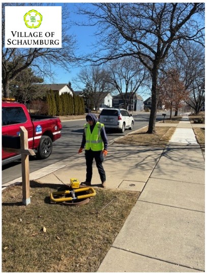 A man in a yellow safety vest stands near a yellow surveying tool on a suburban street in Schaumburg, with a red pickup truck and a white vehicle parked nearby.